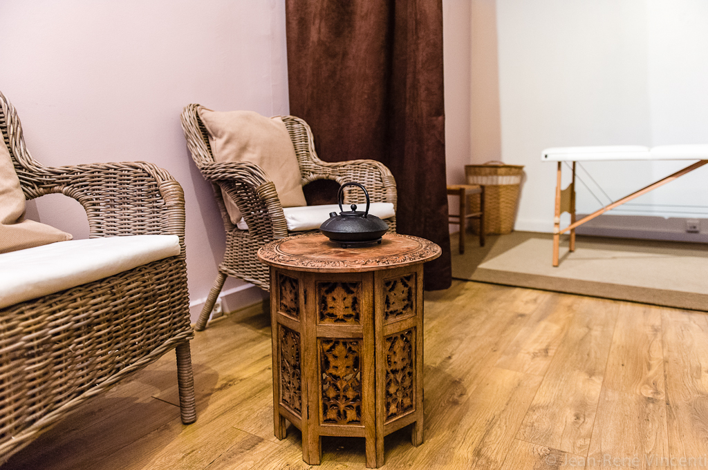 Espace détente, avec fauteuils en rotin, parquet chaleureux en bois, table de massage en arrière plan, et une théeière en fonte posée sur petite table en bois sculpté au premier plan.