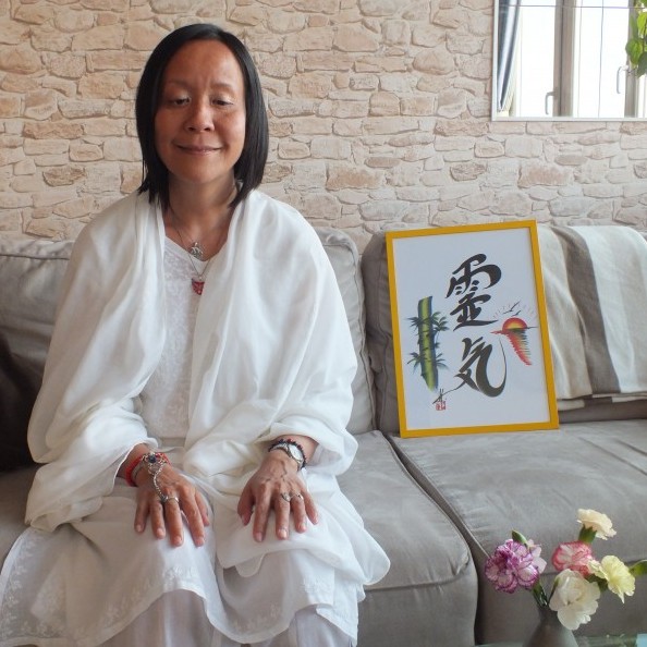 Photo d'Anne Yung, toute de blanc vêtue, assise à côté d'un tableau représentant l'idéogramme du Reiki.